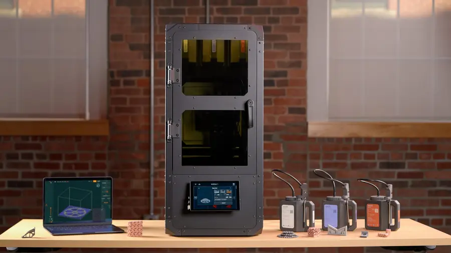 The Mesa 1 printer on a table in front of a brick wall, with a laptop running our slicer to its left, three material cartridges to its right and printed metal parts along the front